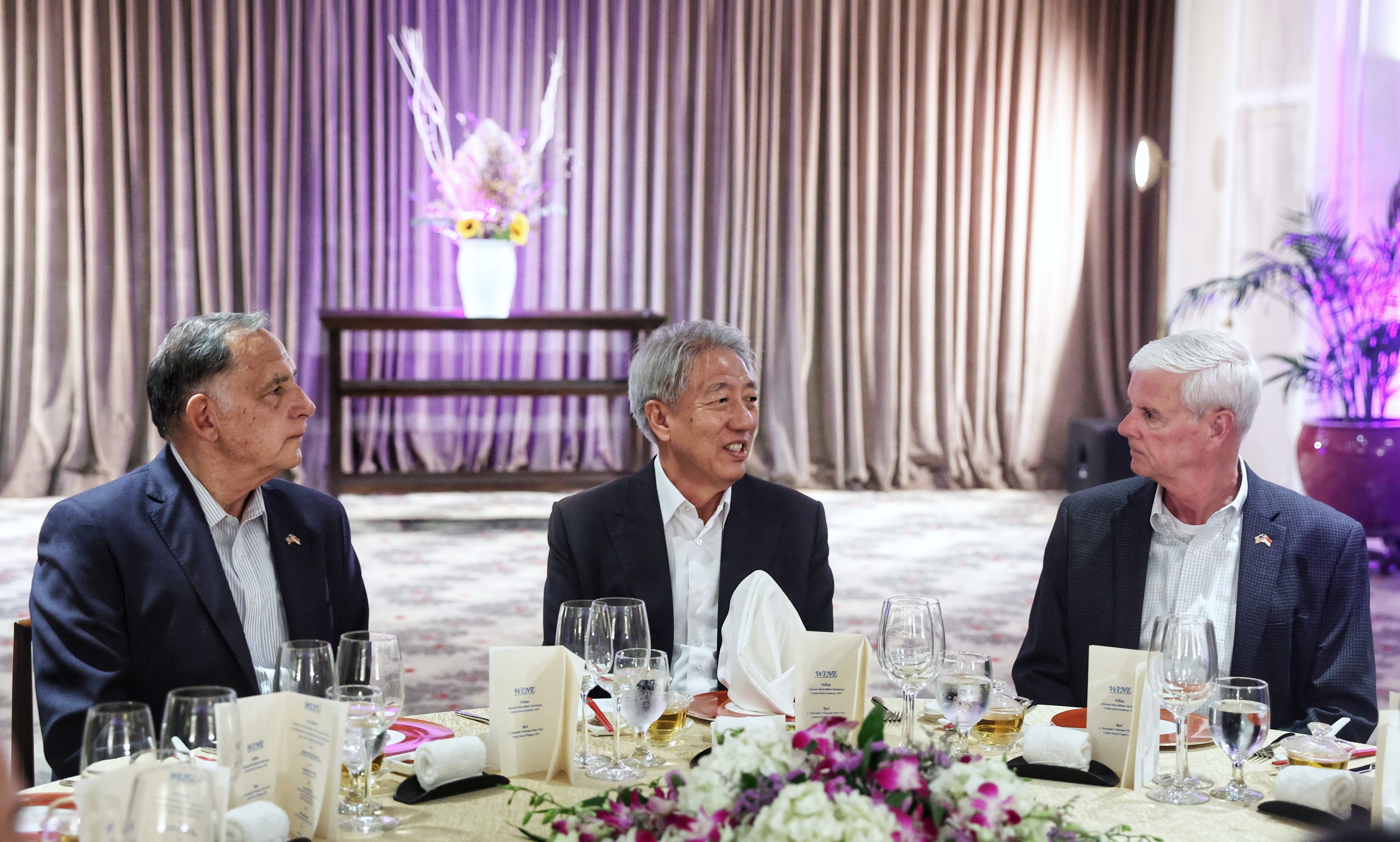 Three men in suits seated at a formal dinner table with floral arrangements.